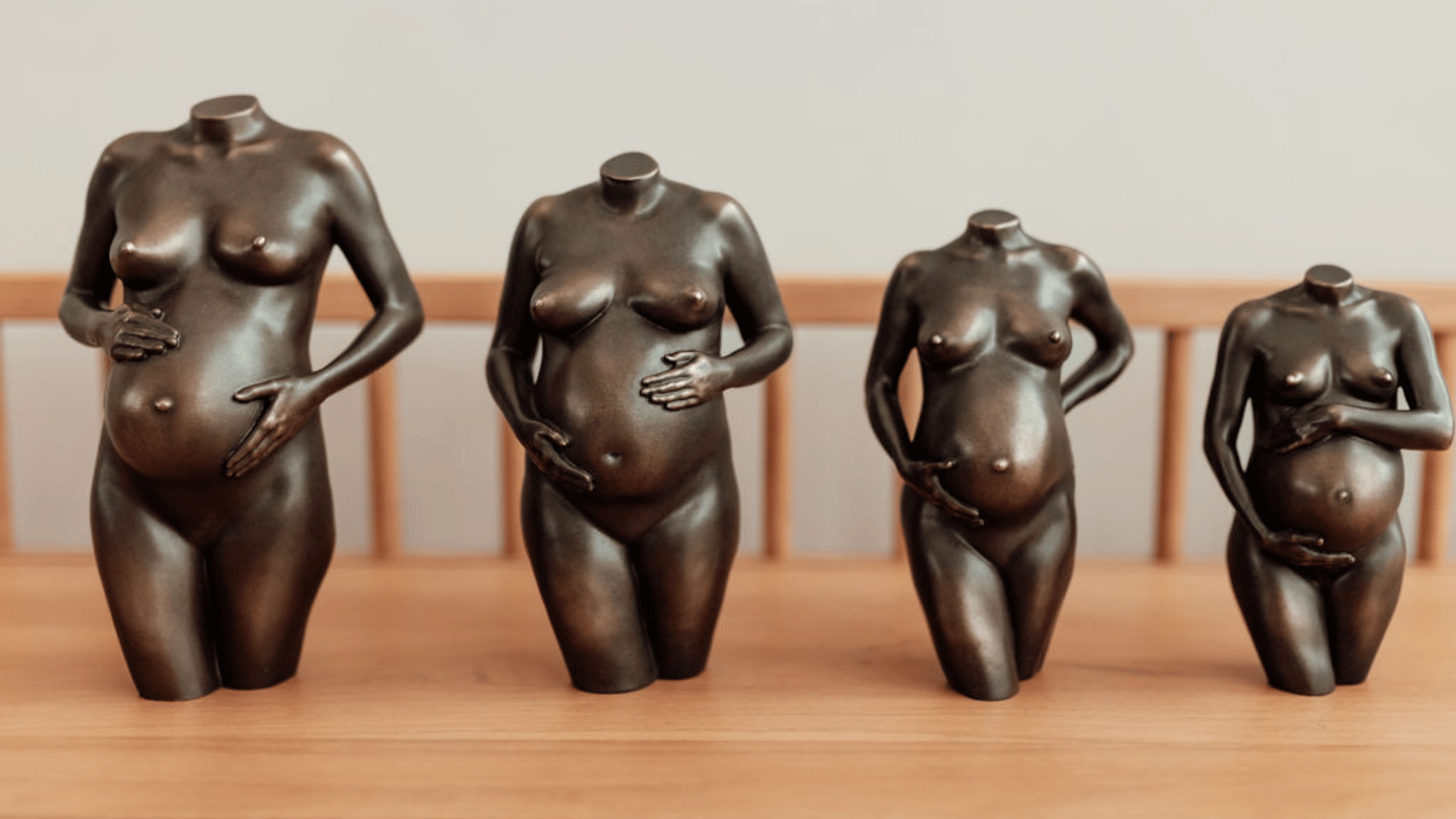Vier bronskleurige zwangerschapsbeeldjes met verschillende handhoudingen naast elkaar op een houten tafel.